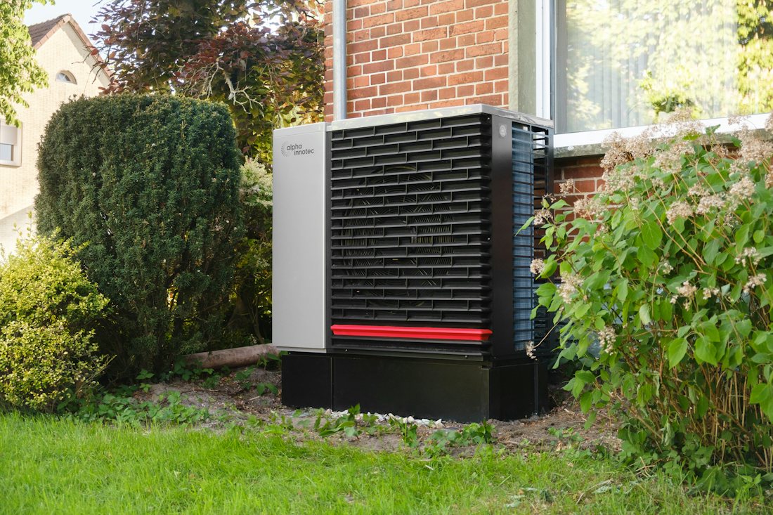 Outdoor heat pump unit beside a brick house, framed by shrubs and lawn